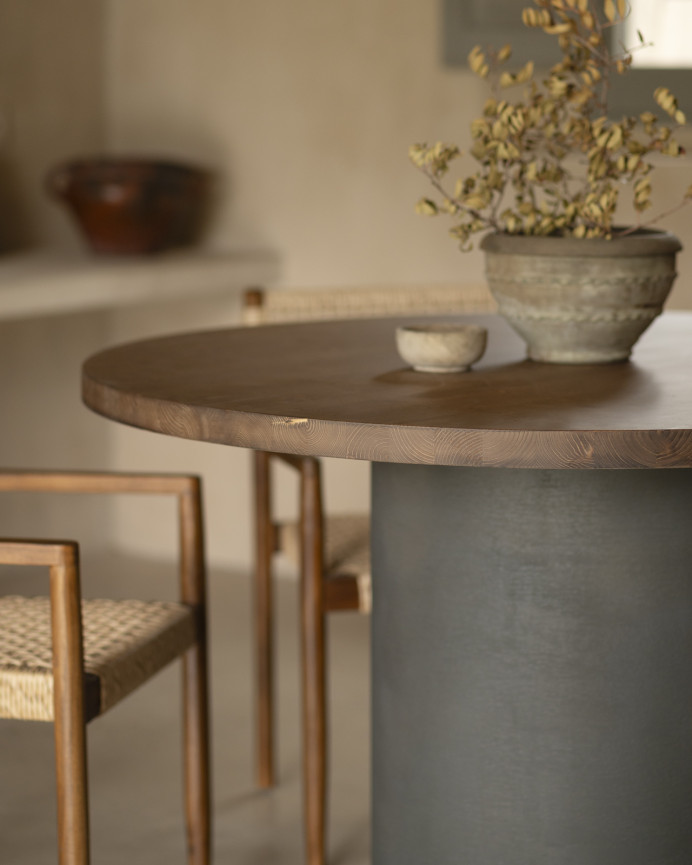Round solid wood dining table in a dark oak tone with microcement legs in a green tone, available in various sizes.